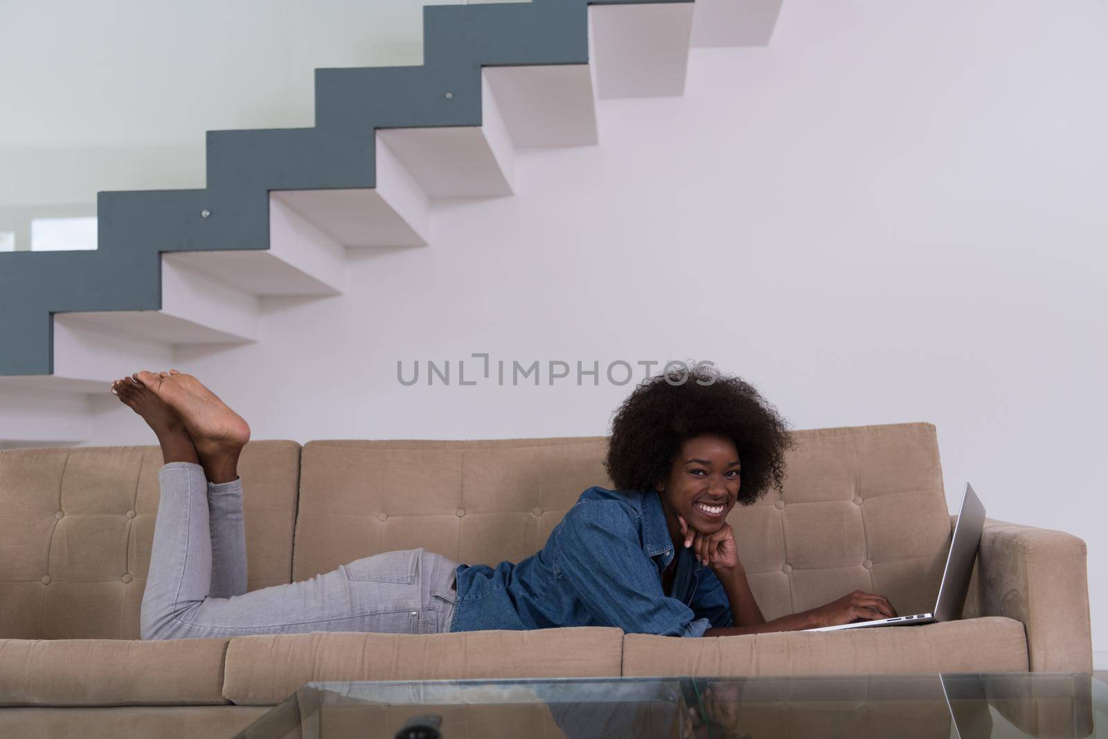Smiling young African American woman using laptop while relaxing on sofa at luxury home