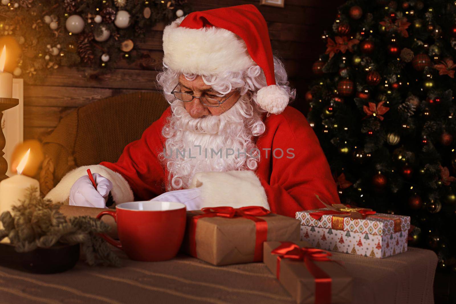Portrait of happy Santa Claus sitting at his room at home near Christmas tree and answering Christmas letters
