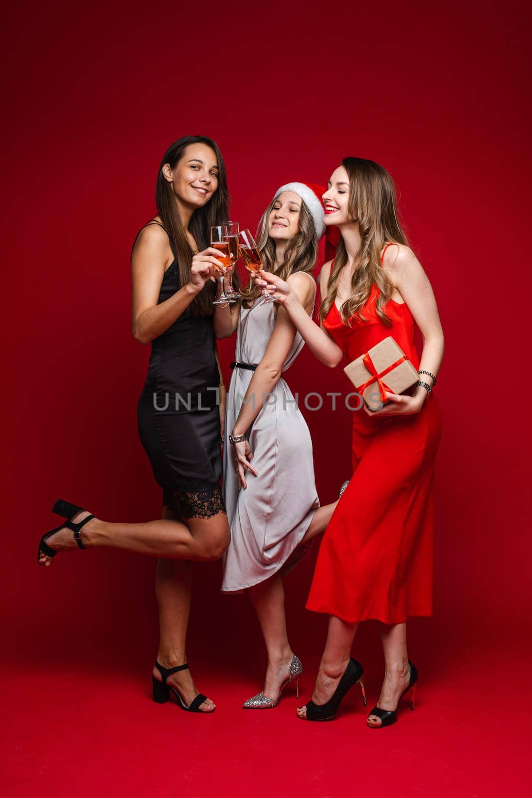 Full length of smiling pretty three girlfriends clinking glasses with champagne, isolated on red background. Party concept