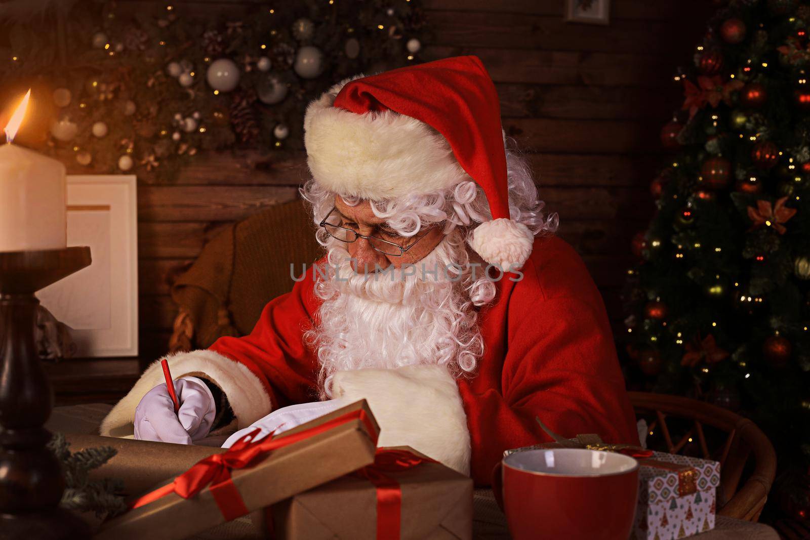 Portrait of happy Santa Claus sitting at his room at home near Christmas tree and answering Christmas letters