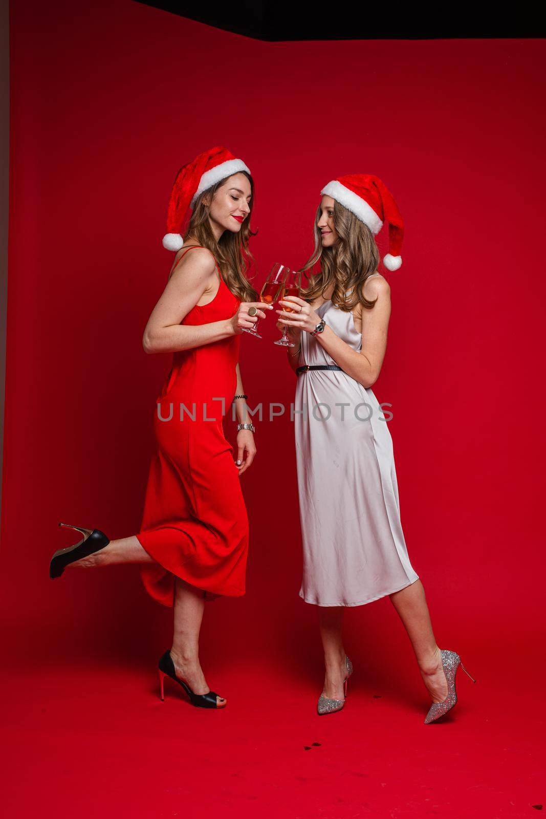 Studio portrait of two attractive girlfriends with long wavy hair in red and white silk dresses posing in Santa hats with glasses of rose wine. Isolated on red background.