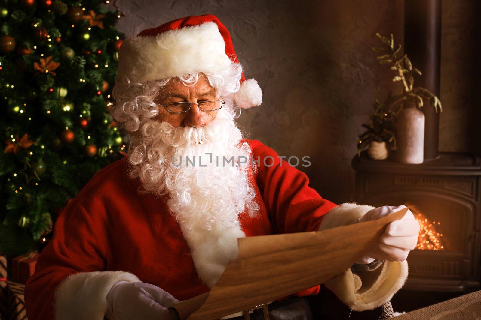 Portrait of happy Santa Claus sitting at his room at home near Christmas tree and reading Christmas letter or wish list