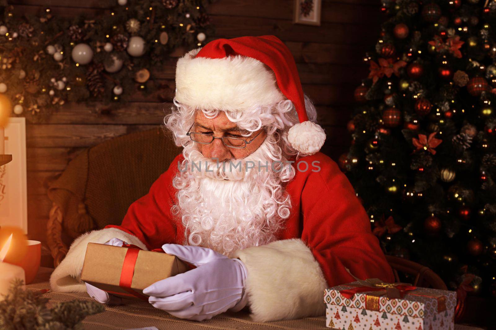 Portrait of happy Santa Claus sitting at his room at home near Christmas tree with gift box