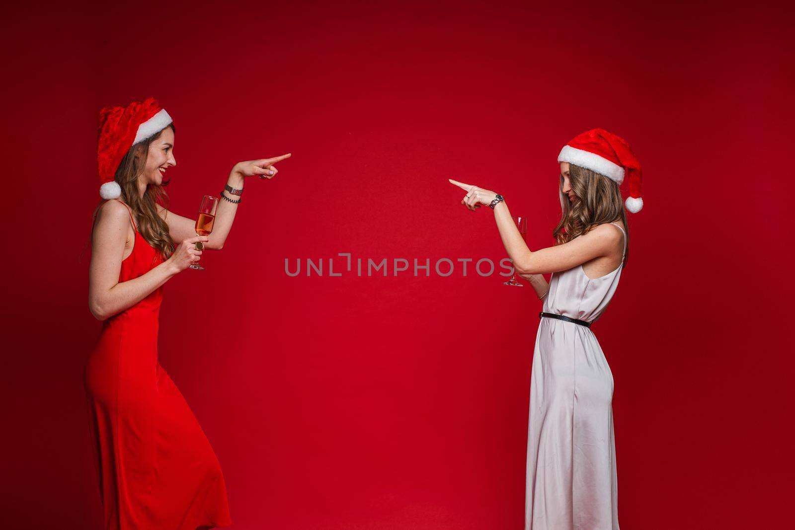 Side view of smiling pretty young women in santa hats pointing fingers at each other, isolated on red background. Party concept