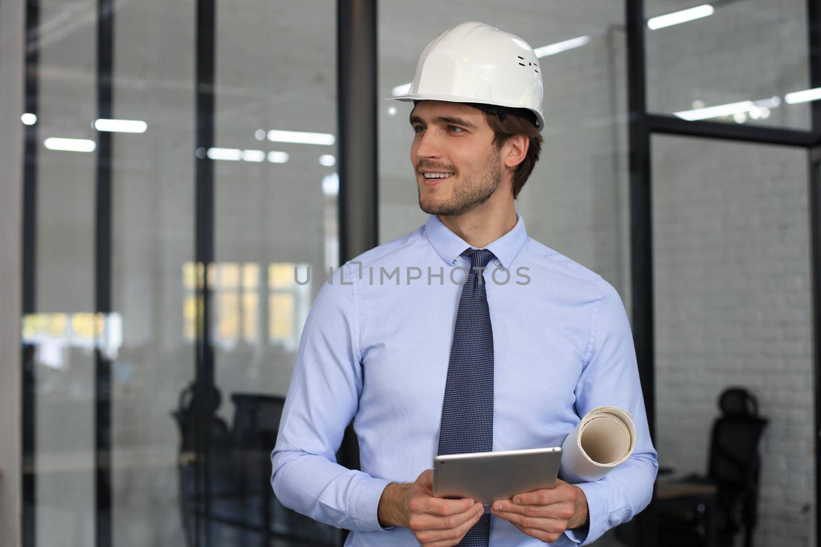 Confident young business man in hardhat holding blueprint and looking away while standing indoors