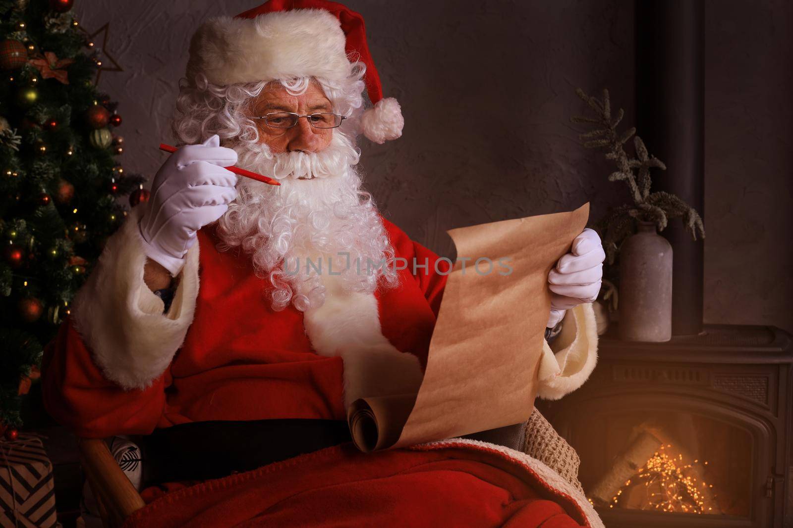 Portrait of happy Santa Claus sitting at his room at home near Christmas tree and reading Christmas letter or wish list