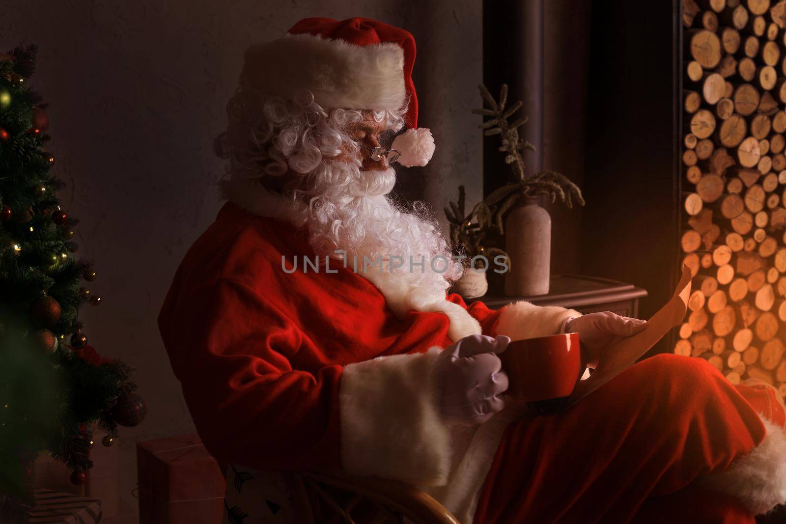 Portrait of happy Santa Claus sitting at his room at home near fireplace and reading Christmas letter or wish list