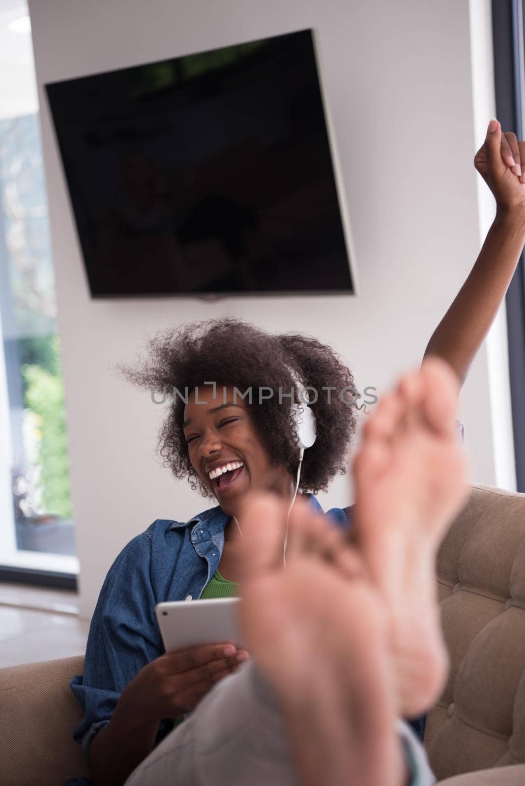 young beautiful African American woman enjoys listening to music with headphones and tablet in your armchair in her luxury home