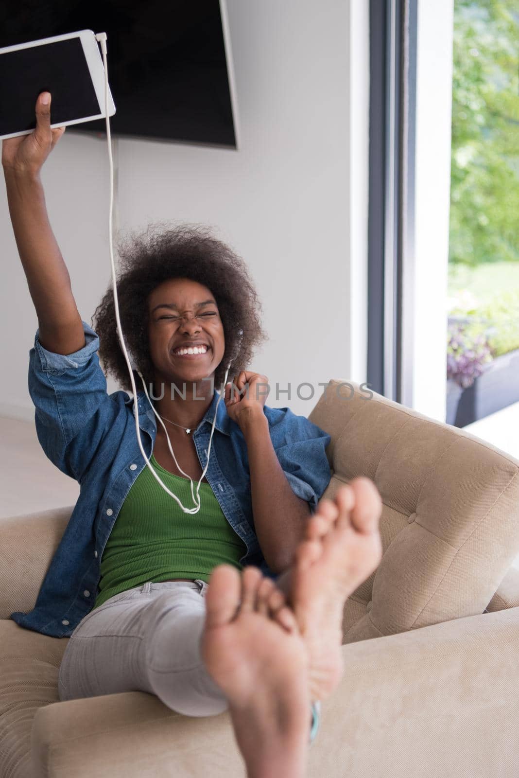 young beautiful African American woman enjoys listening to music with headphones and tablet in your armchair in her luxury home