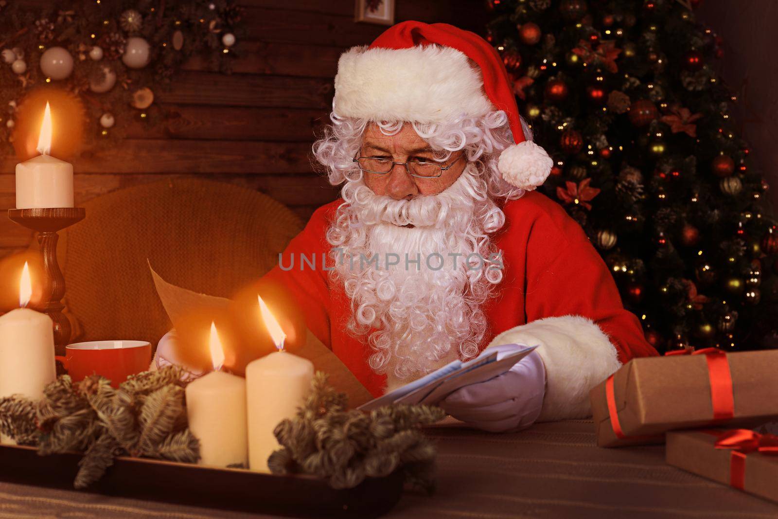 Portrait of happy Santa Claus sitting at his room at home near Christmas tree and reading Christmas letter or wish list