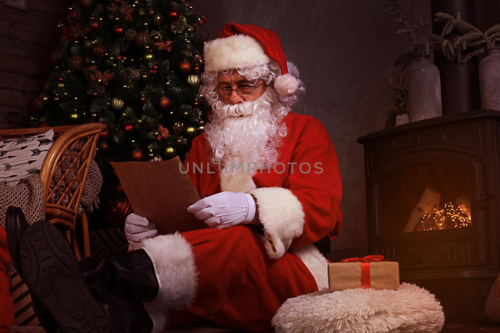 Portrait of happy Santa Claus sitting at his room at home near fireplace and reading Christmas letter or wish list