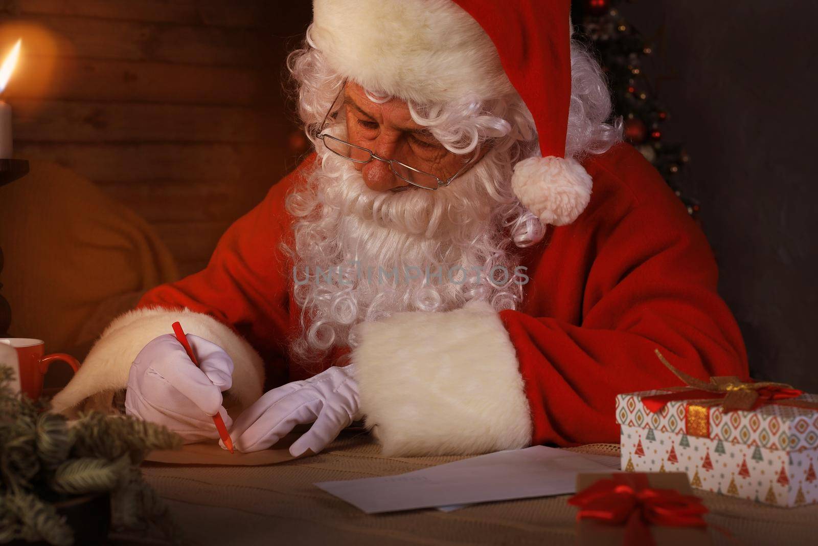 Portrait of happy Santa Claus sitting at his room at home near Christmas tree and answering Christmas letters