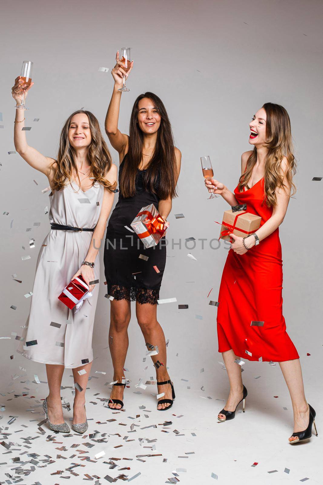 Portrait of jovial carefree beautiful ladies in cocktail dresses with glasses of alcoholic drinks dancing and having fun under falling silver confetti. Girls with drinks holding wrapped gifts.