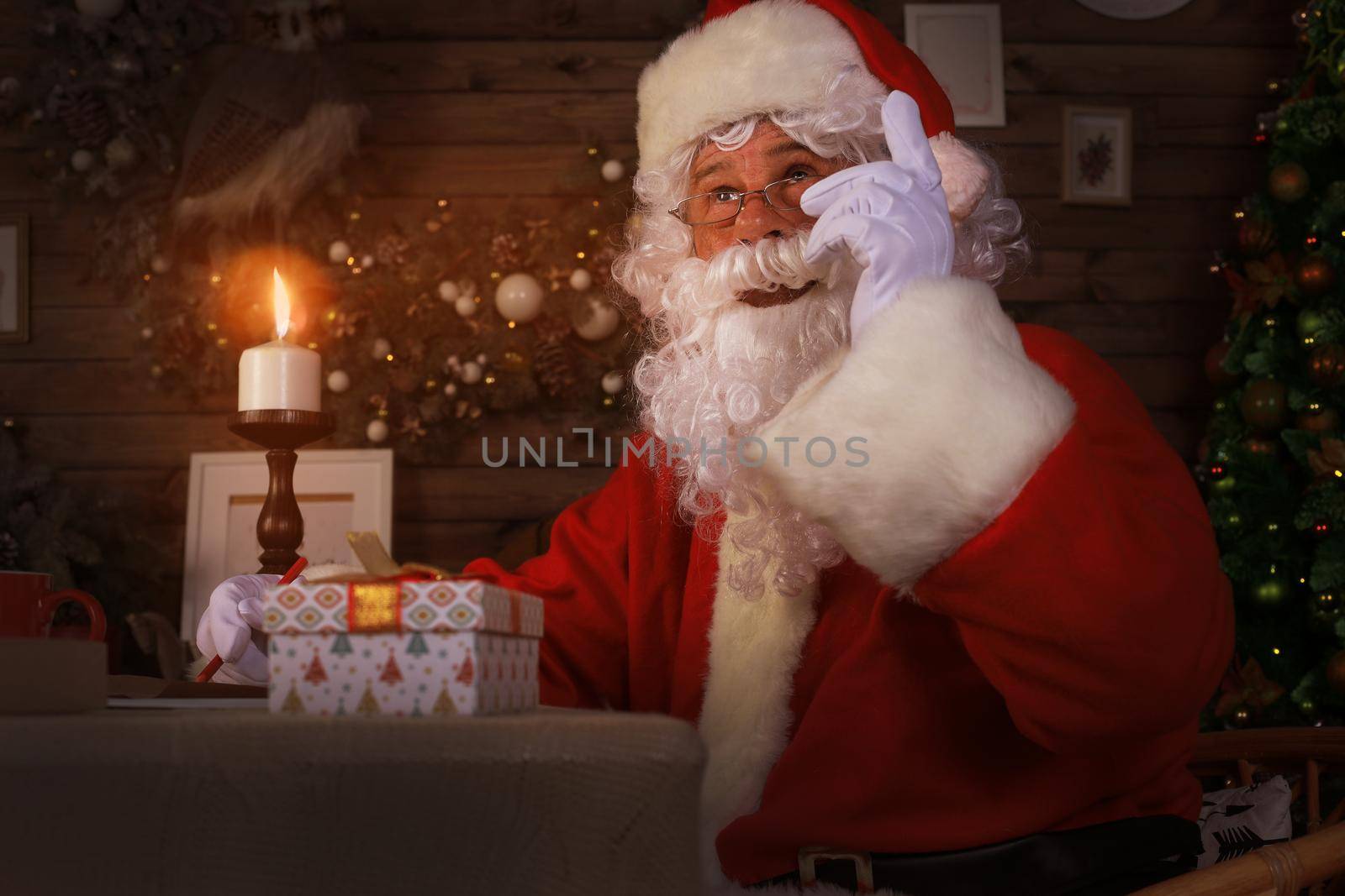 Portrait of happy Santa Claus sitting at his room at home near Christmas tree and answering Christmas letters