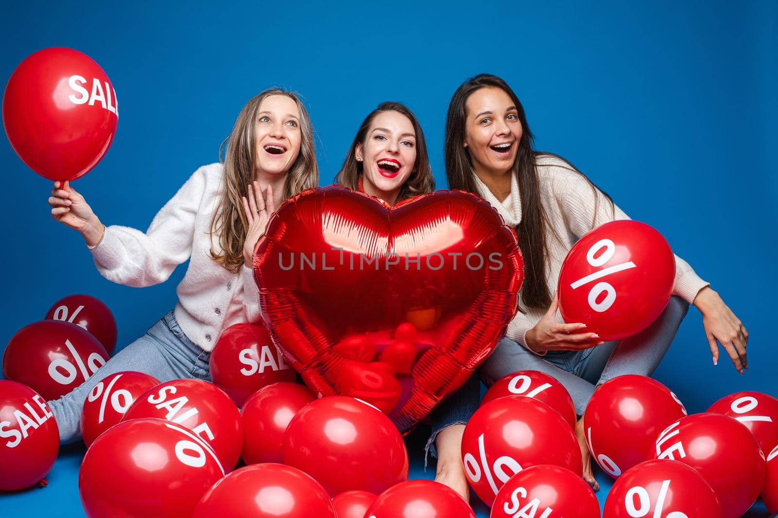 Happy women friends posing with red heart shaped balloon and air balls with percent and sale lettering on blue backdrop by StudioLucky
