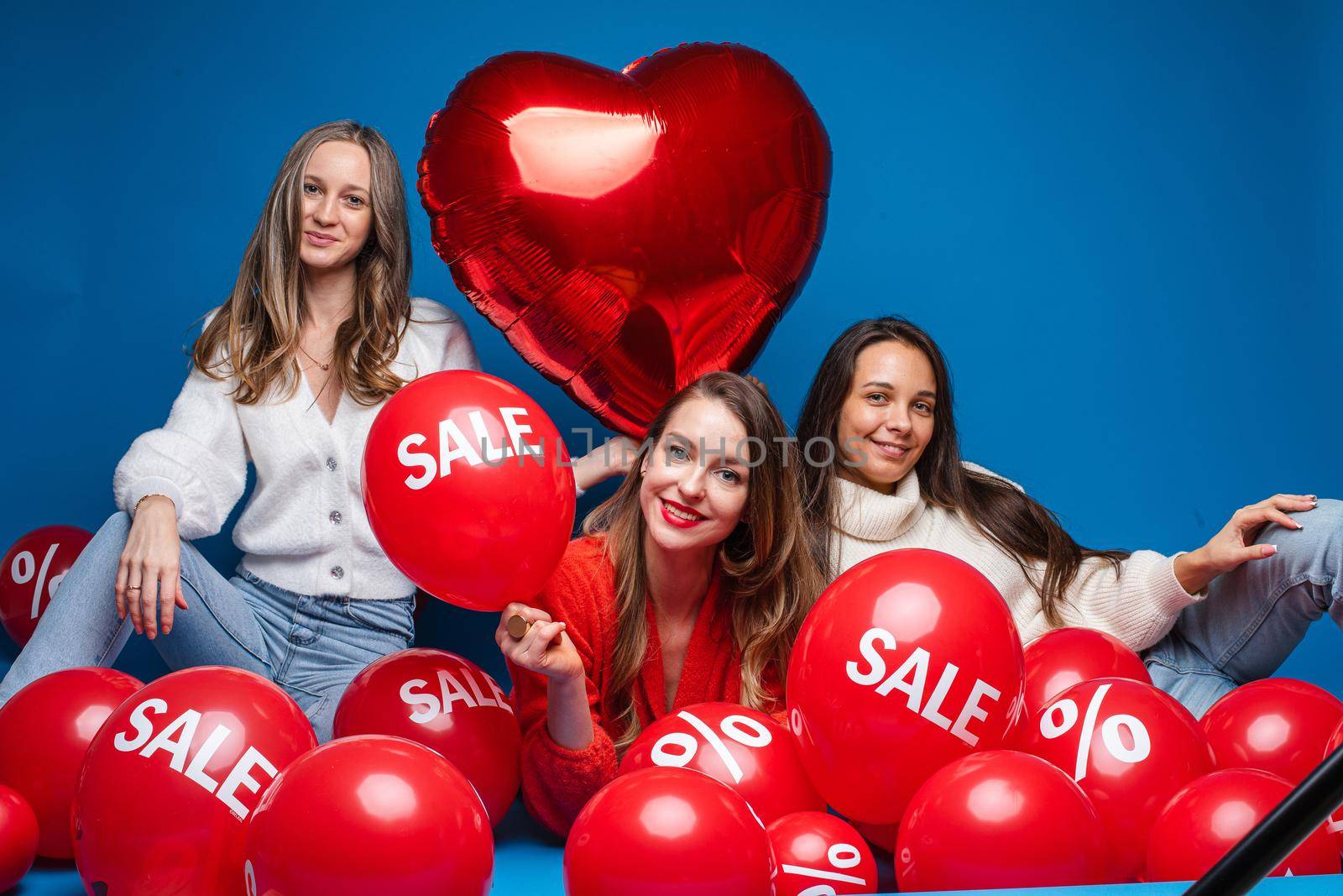 Happy pretty female friends sitting with sale red air balloons, isolated on blue background. Shopping holiday concept