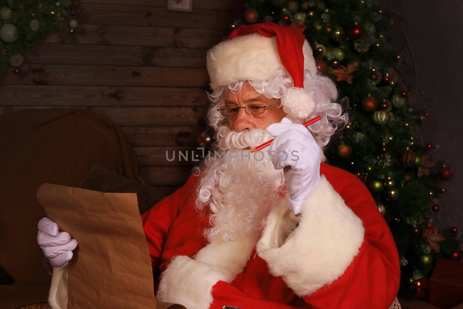 Portrait of happy Santa Claus sitting at his room at home near Christmas tree and reading Christmas letter or wish list
