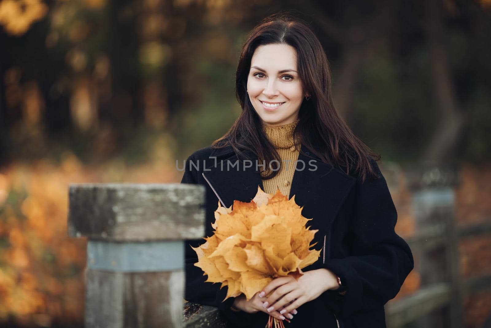 Cheerful young caucasian female with dark hair in brown blouse and black coat smiles outside