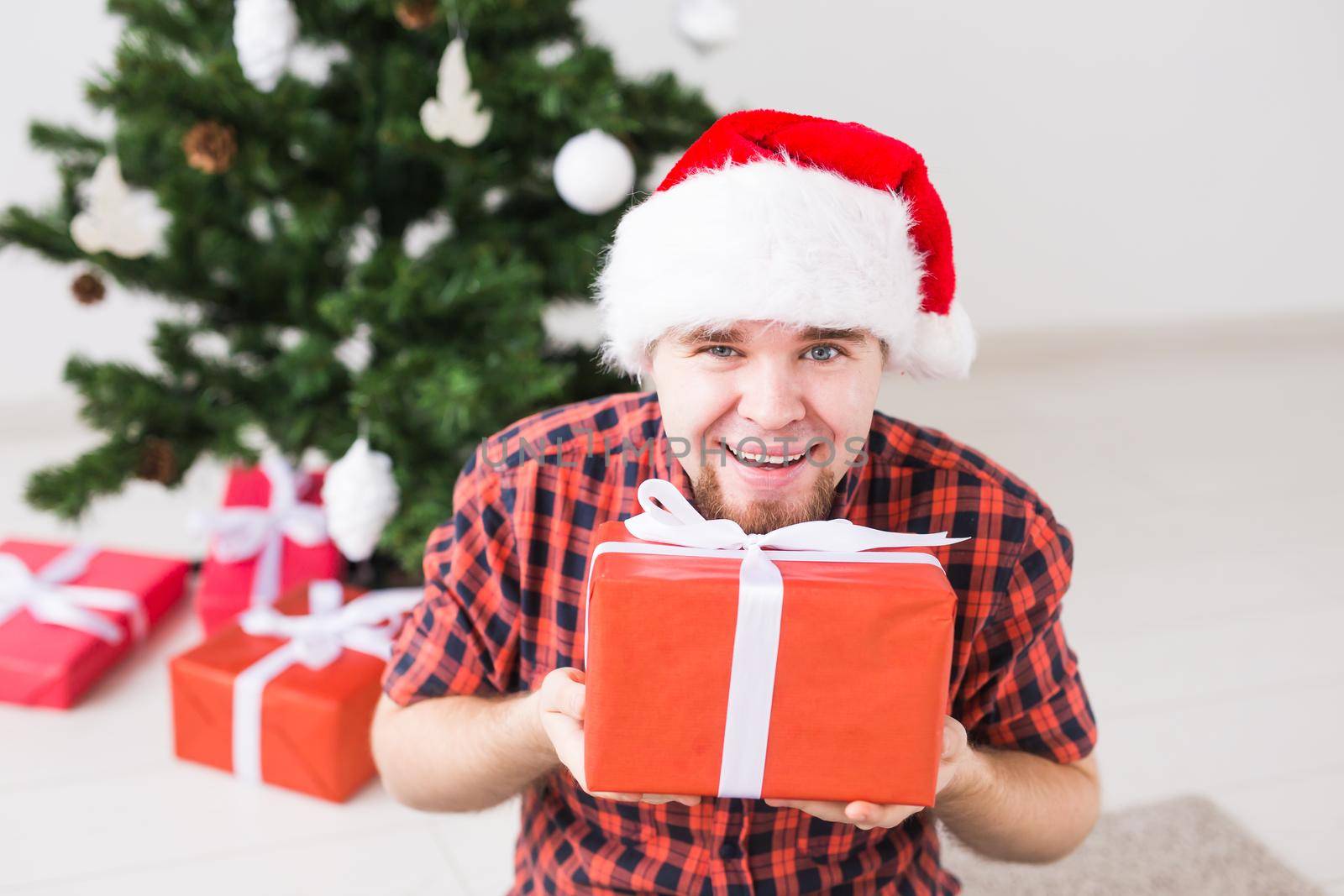 Christmas and holidays concept - Funny man in santa hat holding a gift at home in the living room.