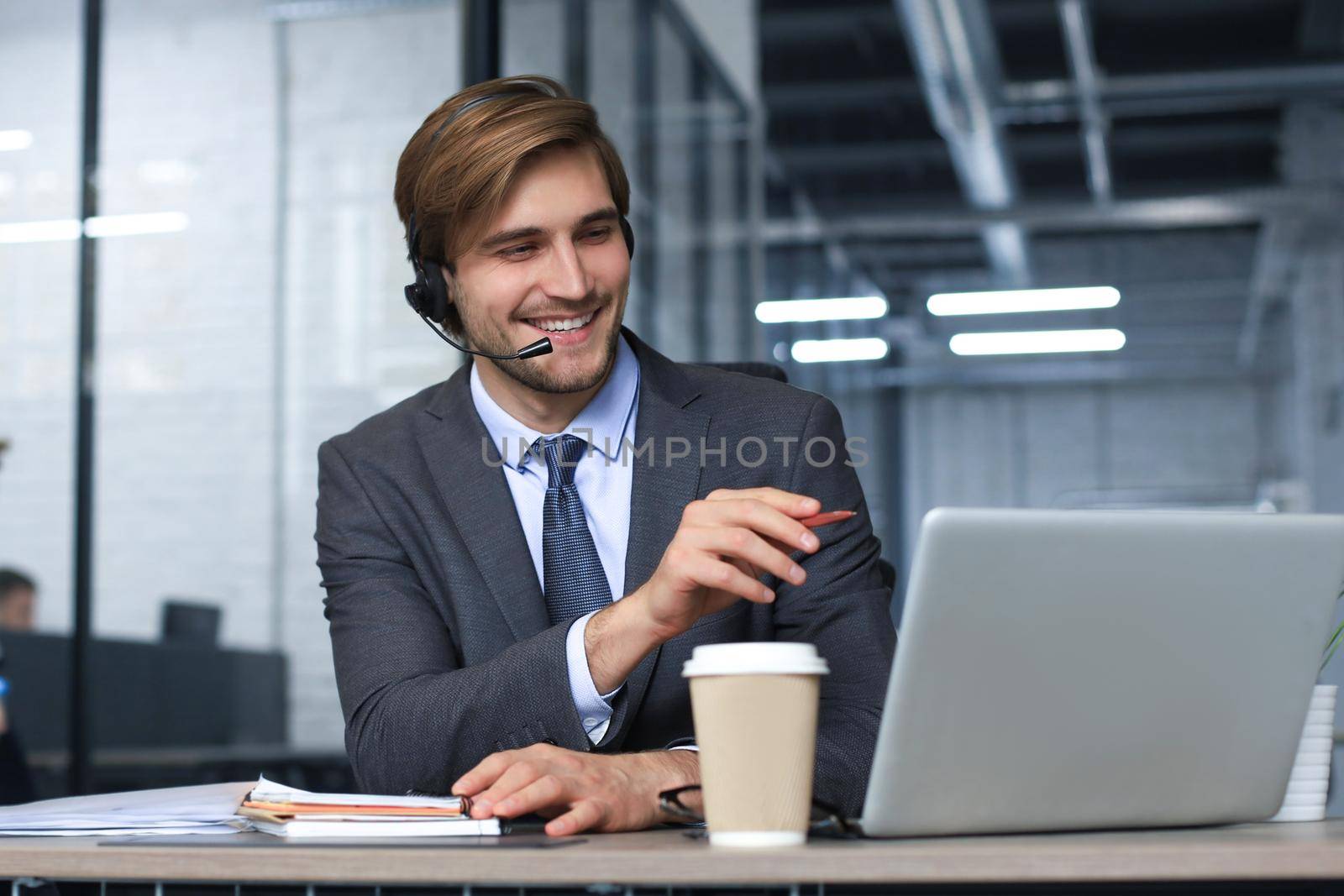 Smiling male call-center operator with headphones sitting at modern office, consulting online information in a laptop, looking up information in a file in order to be of assistance to the client
