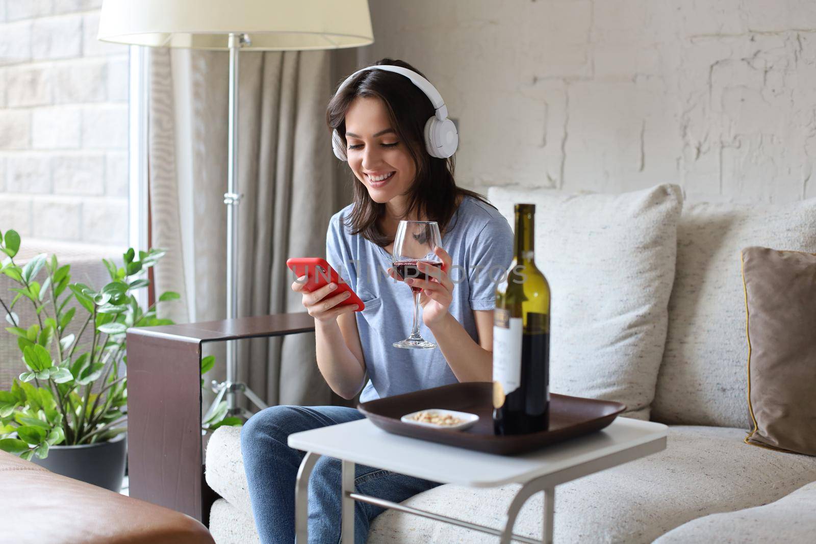 Pretty girl using her smartphone on couch at home in the living room. Listening music, drinking red wine, relaxation after a hard week at work