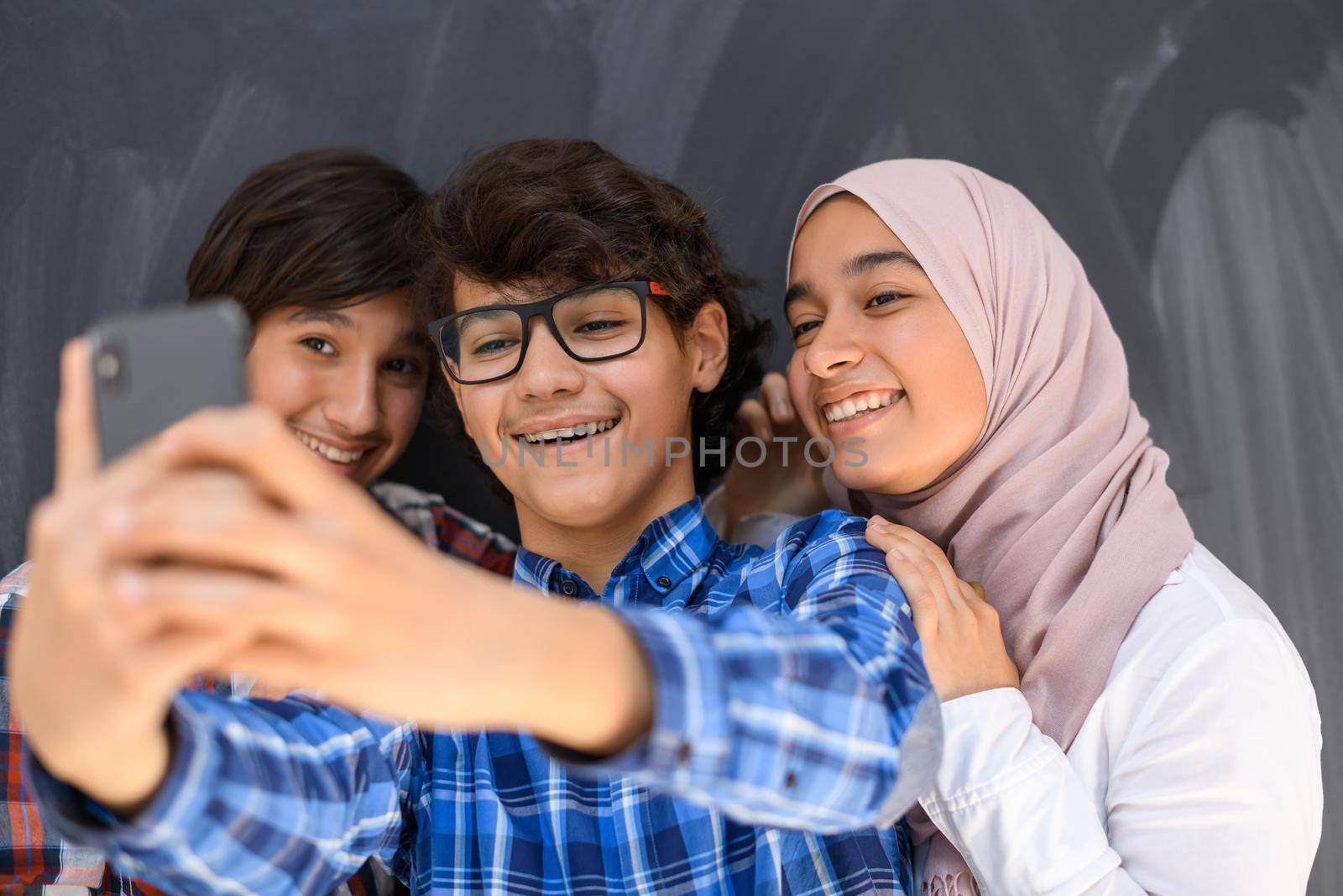 Group of Arab teens taking selfie photos on a smartphone with a black chalkboard in the background. Selective focus. High quality photo
