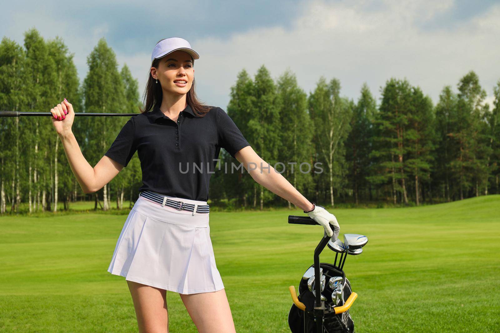 Woman in stylish summer golf outfit walking with bag of drivers on beautiful green course for golf game