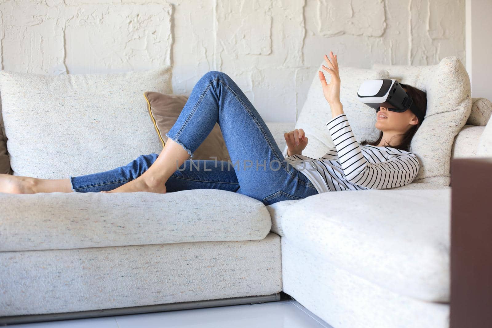 Young casual girl wearing VR headset while lounging on couch at home and looking around