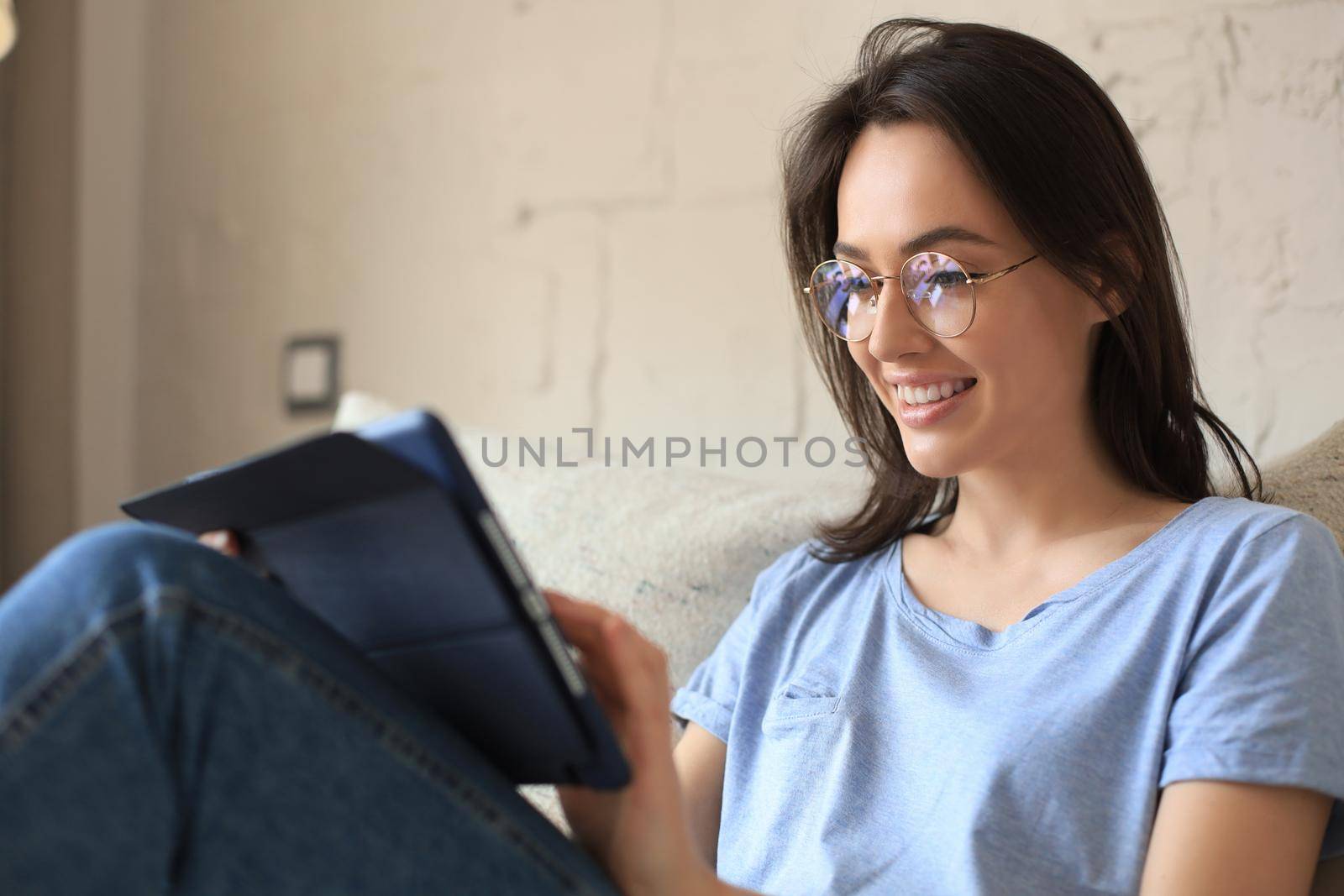 Smiling young woman sitting on sofa with digital tablet and chating with friends