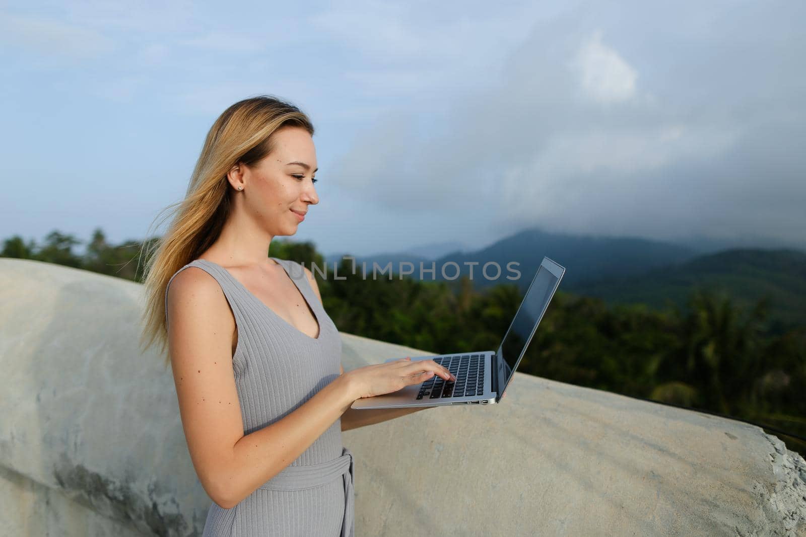 Young woman using laptop in green mountains background, wearing grey shirt. Concept of modern technology, summet vacations and nature.