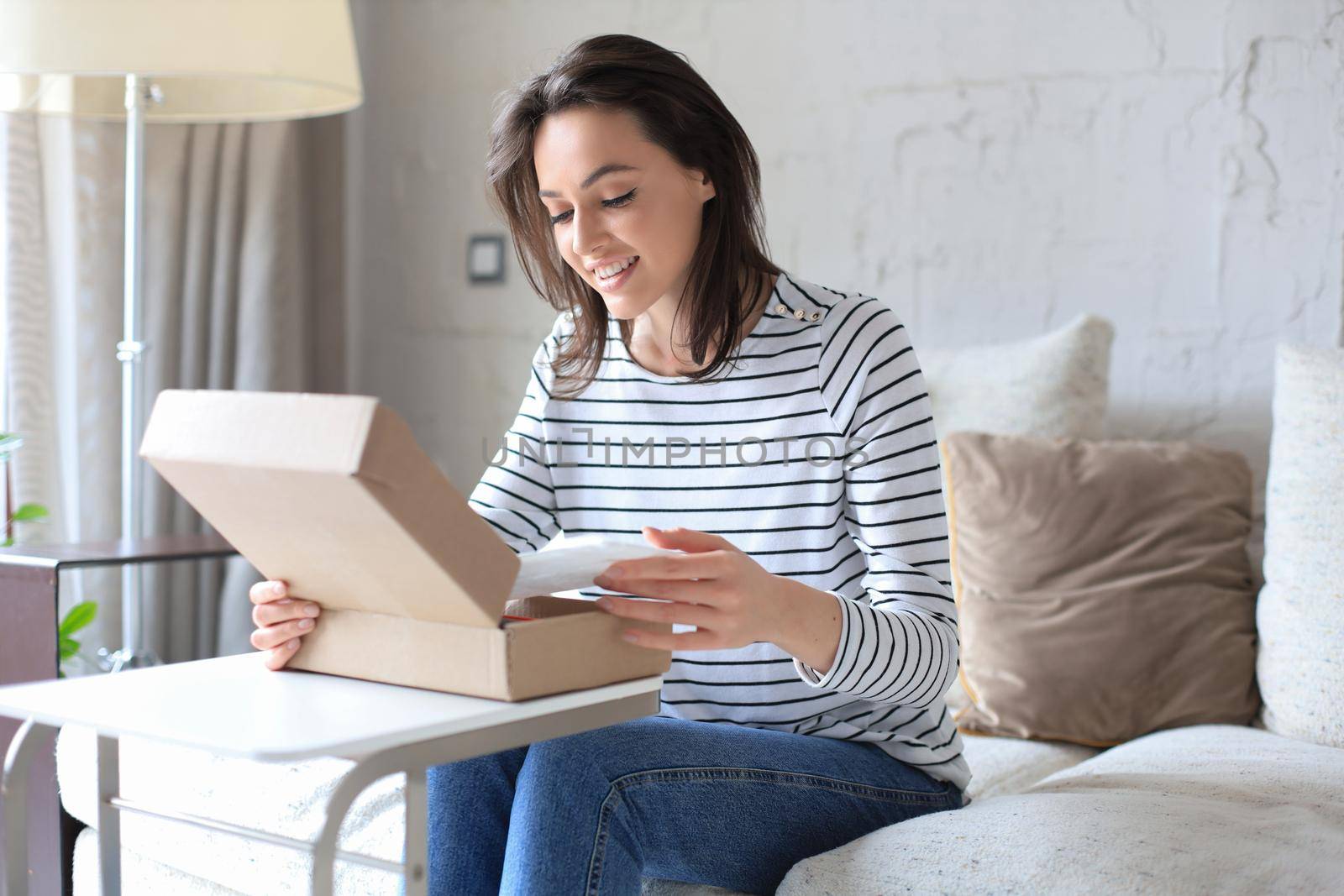 Beautiful young woman is holding cardboard box and unpacking it sitting on sofa at home
