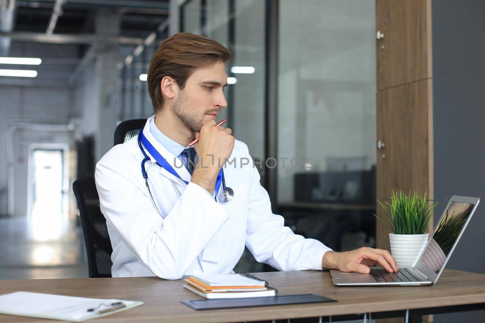 Doctor in white medical uniform with statoscope reading personal file of patient on tablet and writes data to laptop