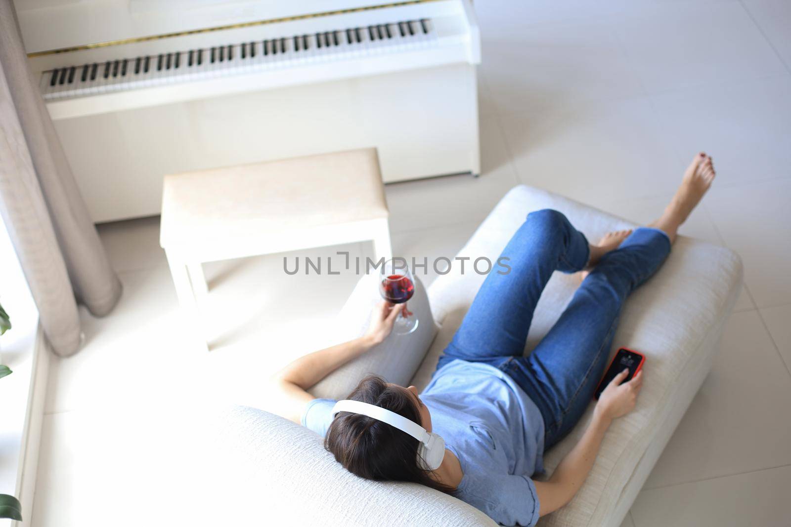 Beautiful brunette woman in earphones sitting on sofa listening to music with closed eyes, drinking red wine