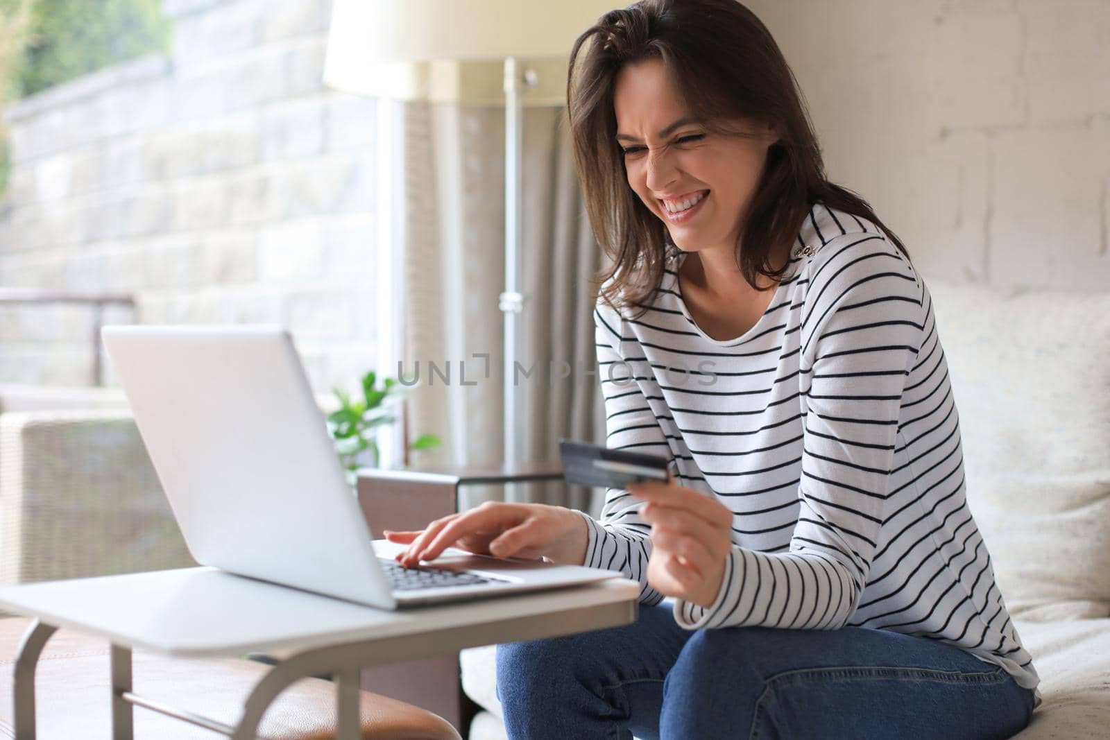 Happy natural brunette using credit card and laptop in the living room