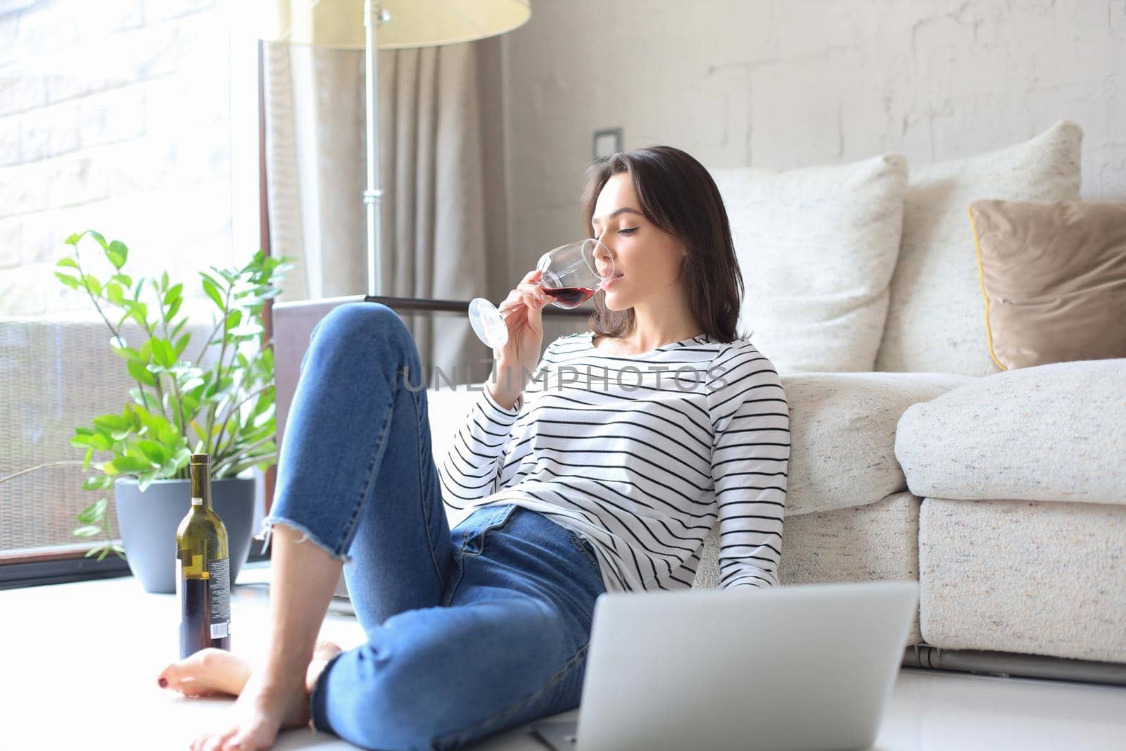 Smiling young woman sitting on floor with laptop computer and chating with friends, drinking wine
