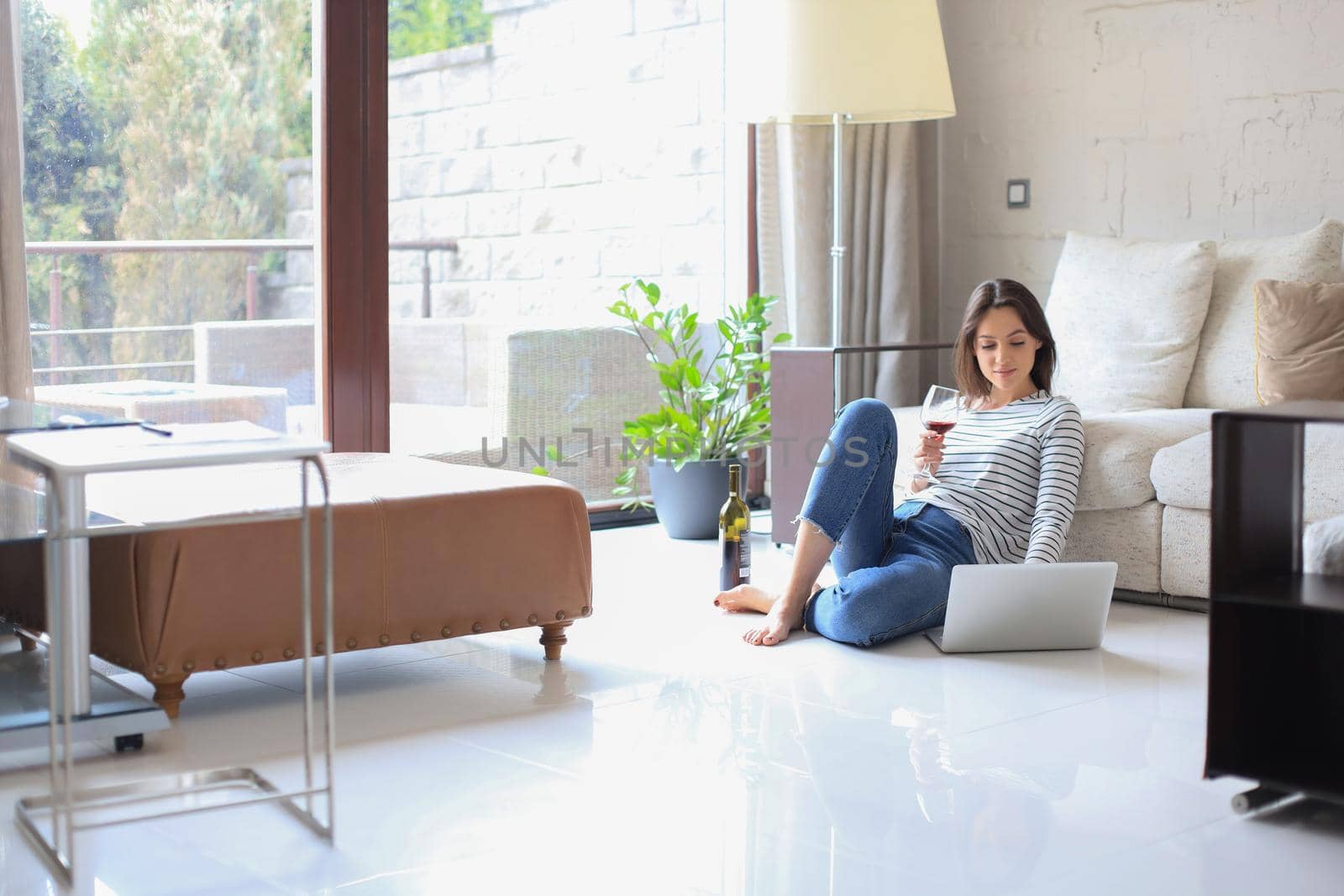 Smiling young woman sitting on floor with laptop computer and chating with friends, drinking wine