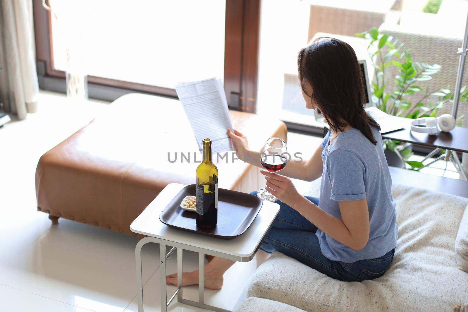 Successful young beautiful woman sitting on a sofa in the living room, drinking red wine, reading documents