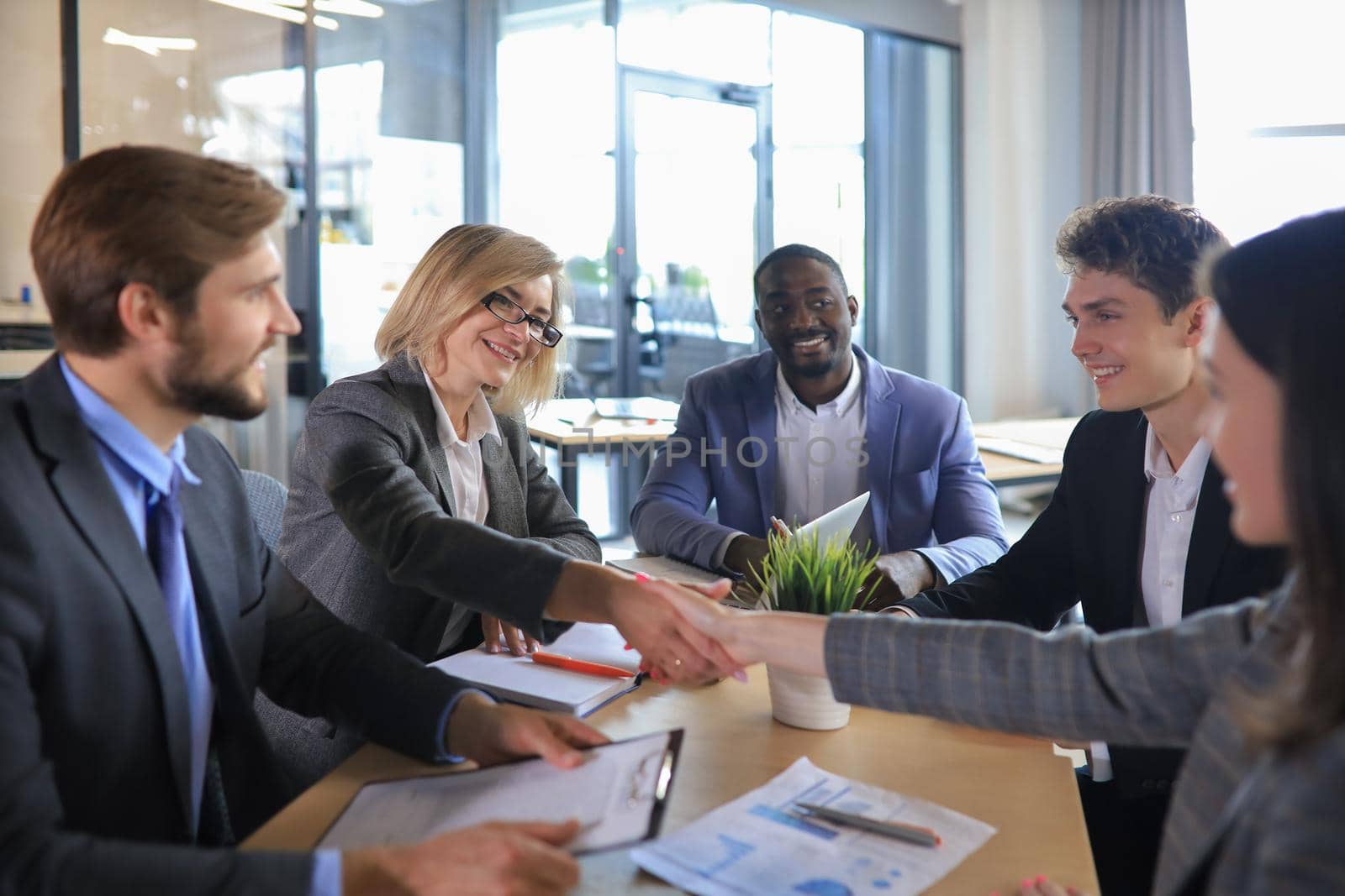 Two women shaking hands and looking at each other with smile while their coworkers sitting at the business meeting