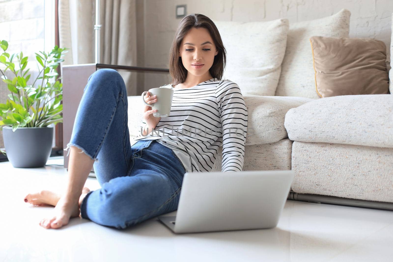 Pretty woman working with laptop while sitting on floor at home and drinking coffee
