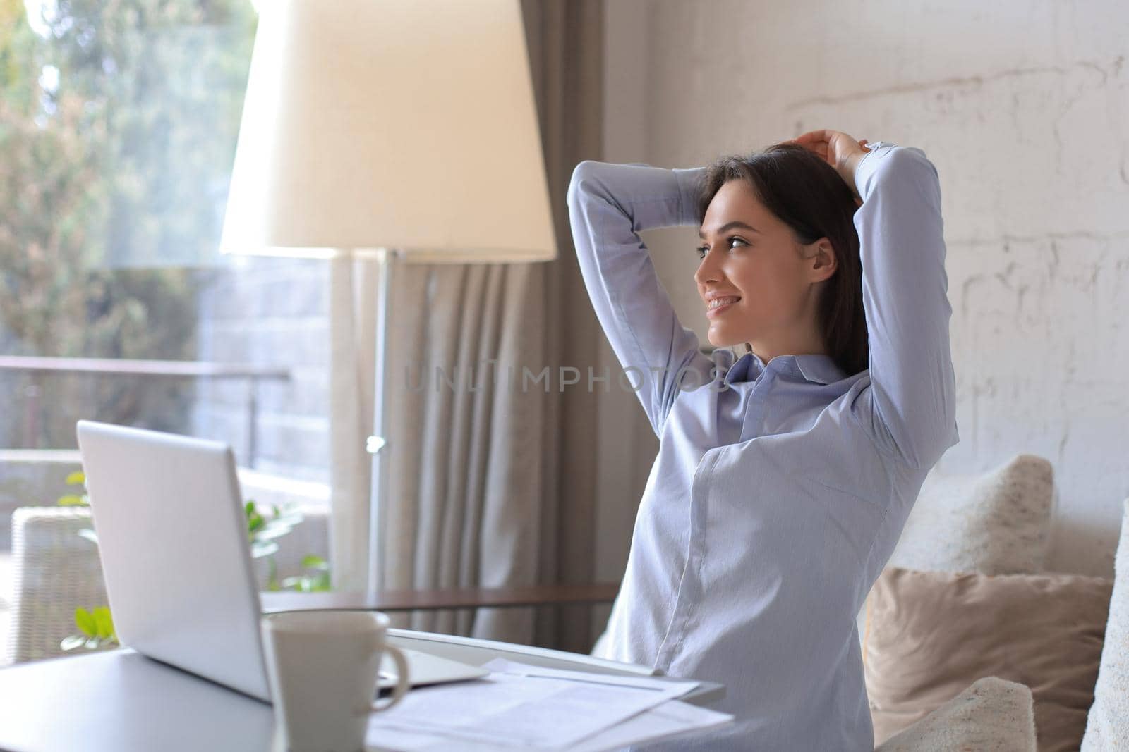 Young woman crossed hands behind head, enjoying break time at home. Peaceful carefree business woman resting at table with computer, looking aside, dreaming of future