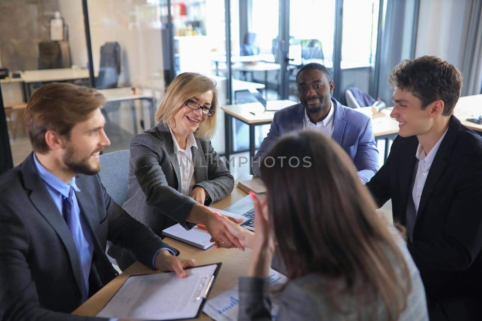 Two women shaking hands and looking at each other with smile while their coworkers sitting at the business meeting