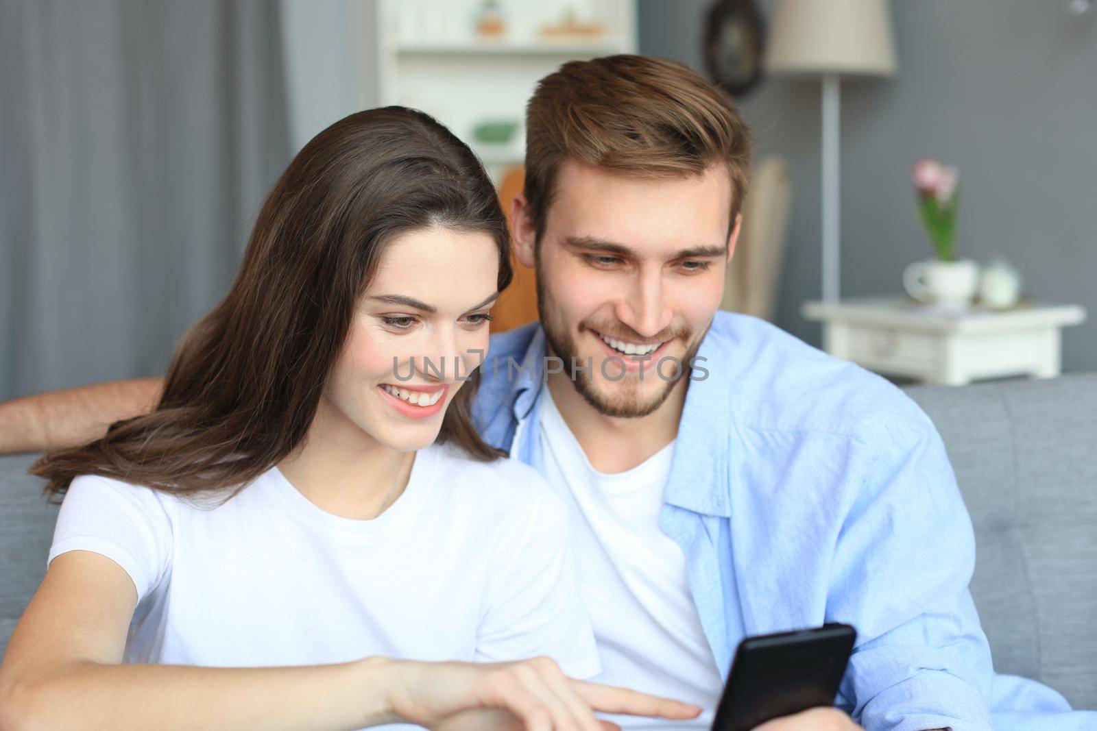 Young couple watching online content in a smart phone sitting on a sofa at home in the living room. by tsyhun