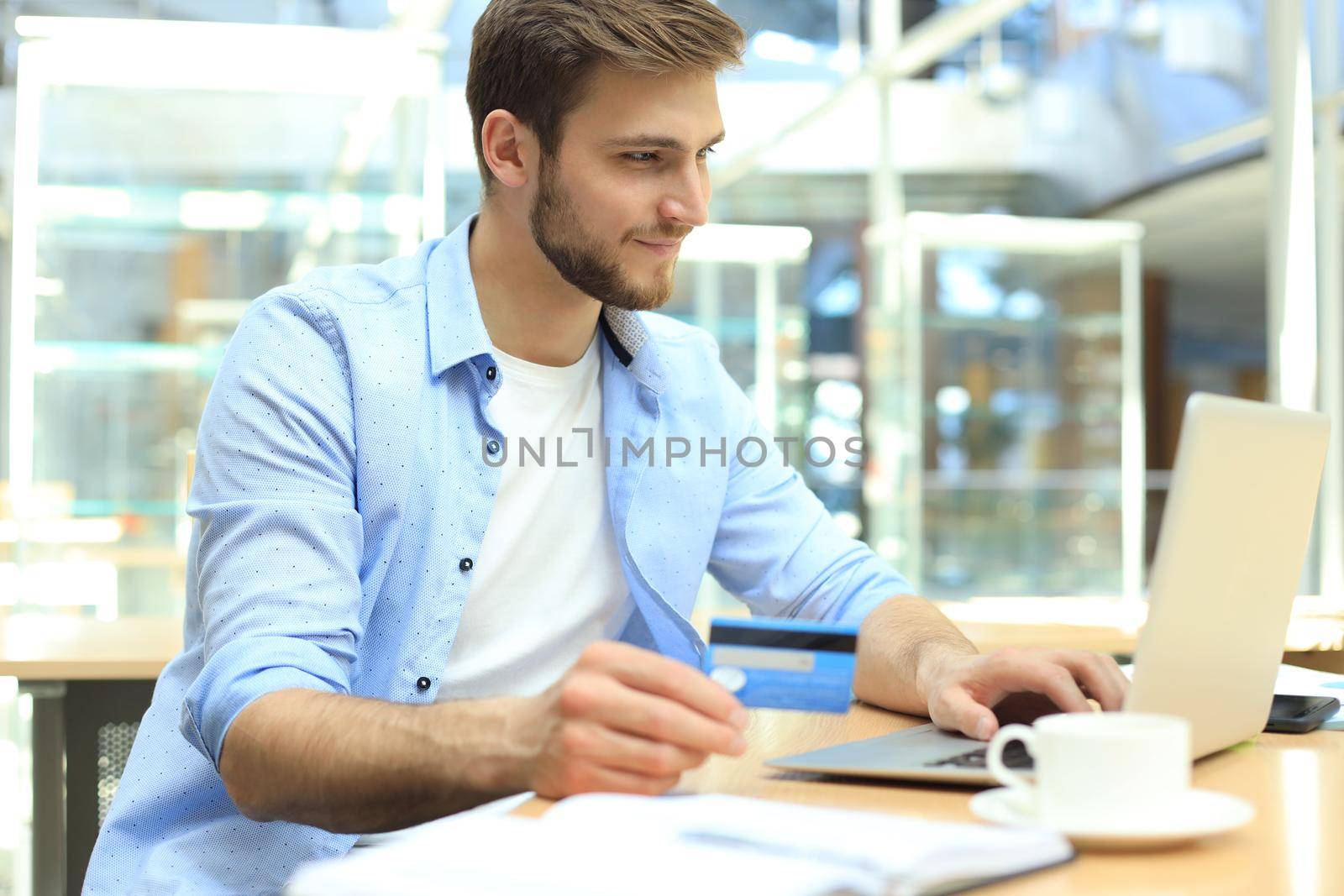 Smiling man sitting in office and pays by credit card with his laptop