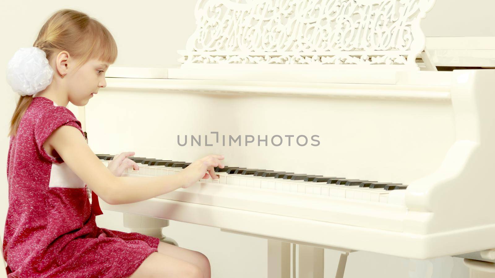 Beautiful little blonde girl with short bangs and pigtails on her head in a good mood.She sits opposite the white piano and tries to play it.