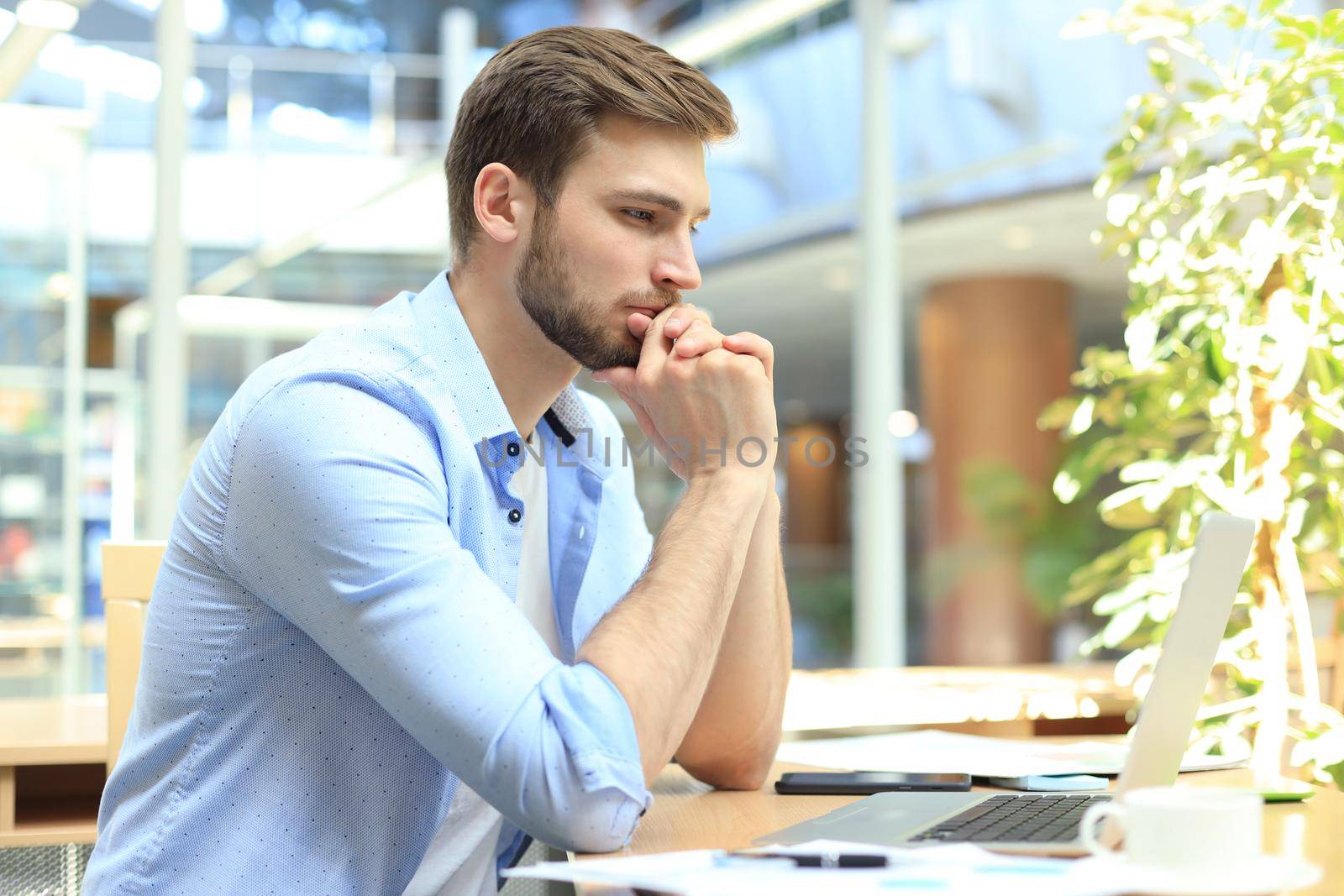 Young man using a laptop sitting thinking at his desk as he reads information on the screen