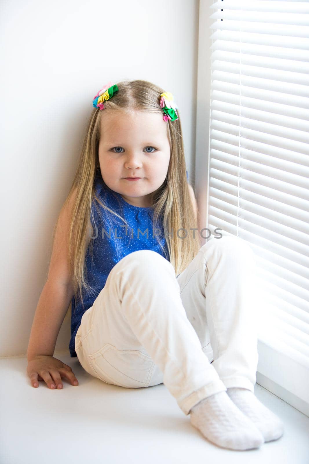 Cute little girl with long blond hair below the shoulders. Blue t-shirt and white jeans sitting on the windowsill
