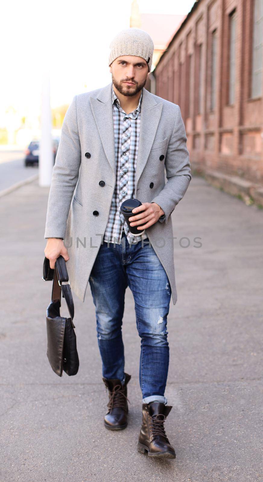 City life. Stylish young man in grey coat and hat walking on the street in the city