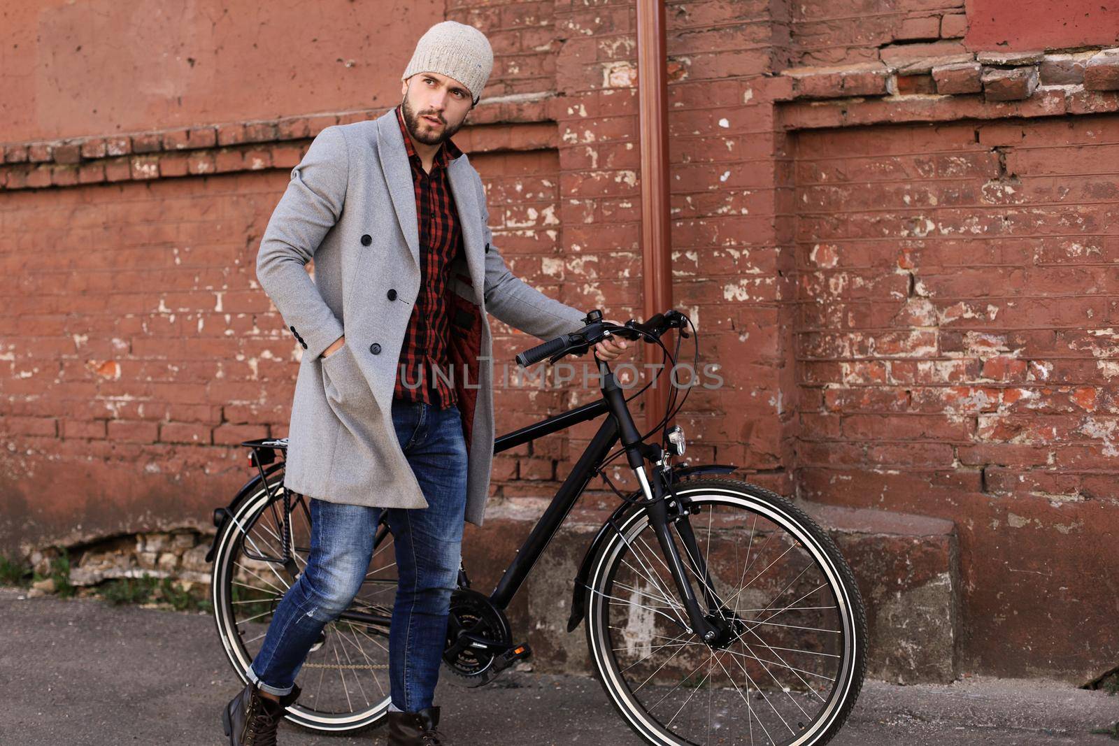 Handsome young man in grey coat and hat pushing his bike down a street in the city