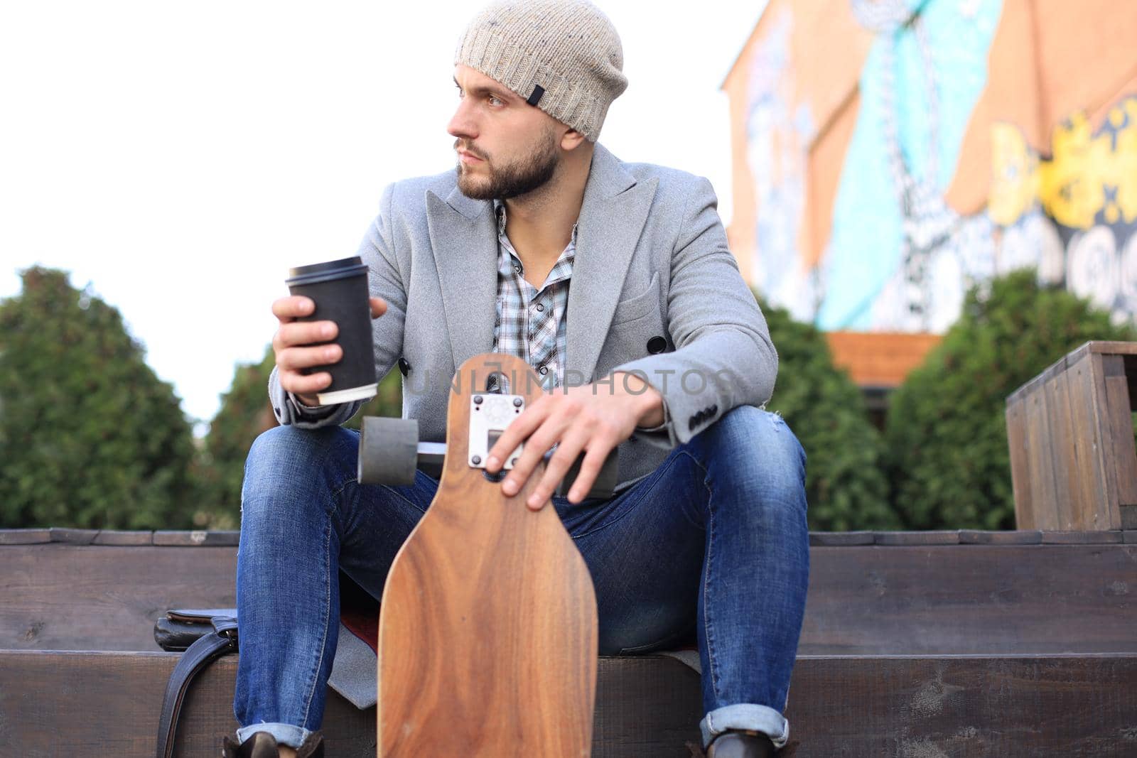 Handsome young man in grey coat and hat, resting, sitting with longboard drinking coffee. Urban skateboarding concept