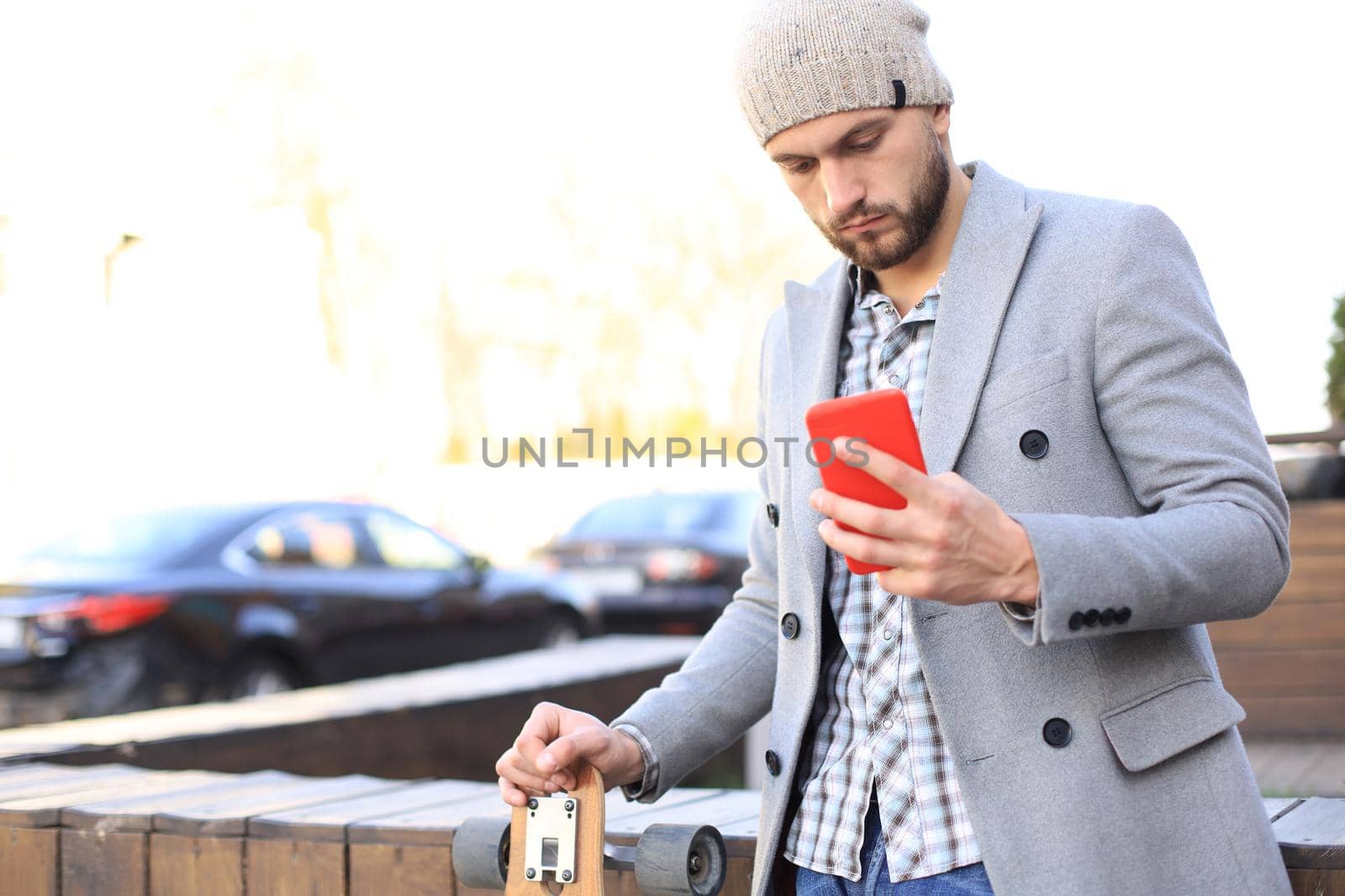 Handsome young man in grey coat and hat using smartphone, resting, standing with longboard. Urban skateboarding concept